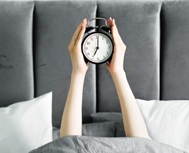 Hands holding an alarm clock above a bed in the morning