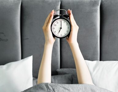 Hands holding an alarm clock above a bed in the morning