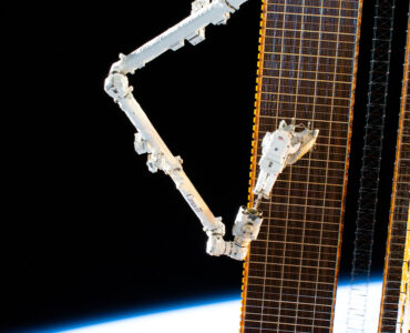 Astronaut Woody Hoburg carries a rolled-up solar array while riding the Canadarm2 robotic arm outside the International Space Station